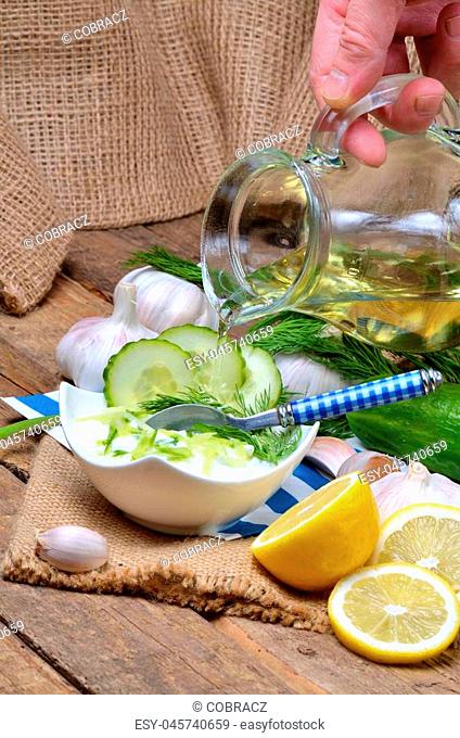 Man pours oil into Tzatziki - traditional Greek dressing or dip sauce, garlic, lemon, dill, cucumber, jug with oil, blue spoon and decoration in background -...