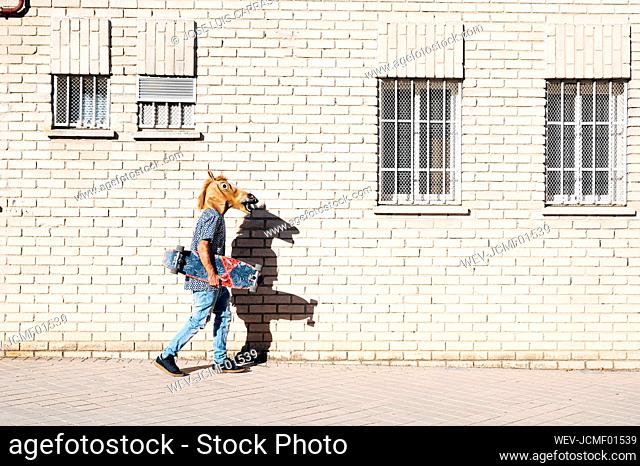 Man walking while holding skateboard wearing horse mask on footpath in city during sunny day
