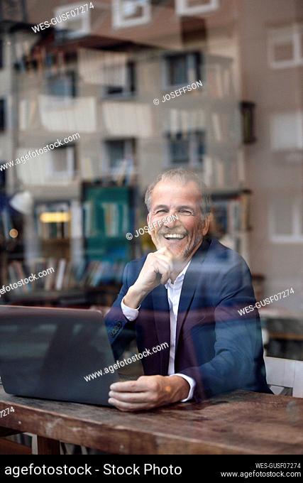 Cheerful businessman with laptop seen through glass window at cafe