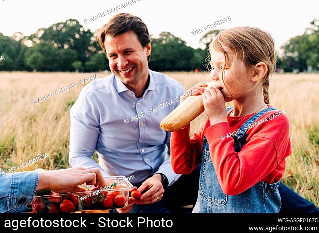 Girl eating loaf of bread at picnic with family in park