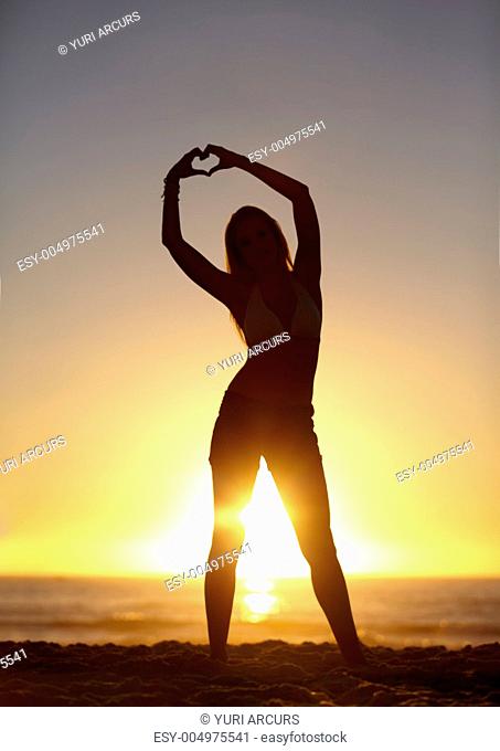 An attractive young woman standing on the beach making heart symbols with her hands