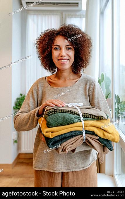 Happy young fashion designer with folded clothes standing in workshop