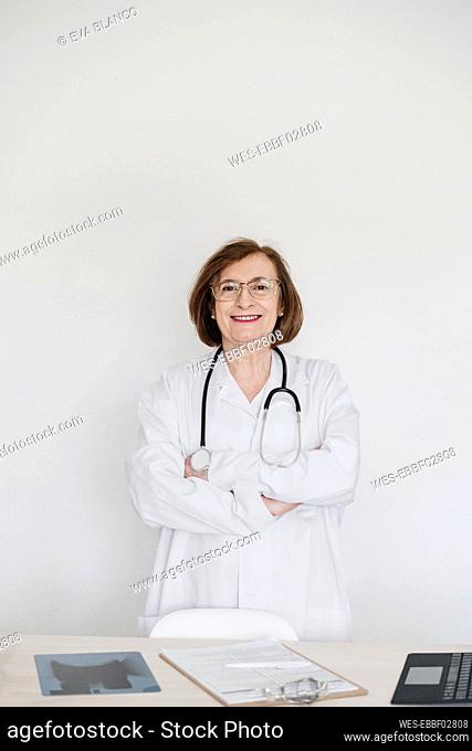 Smiling female doctor with arms crossed standing in front of desk at hospital