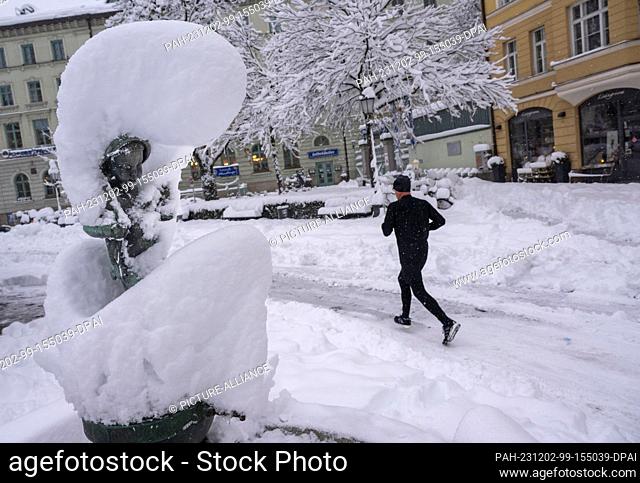 02 December 2023, Bavaria, Munich: A jogger runs along a snow-covered road past a fountain covered in a thick layer of snow
