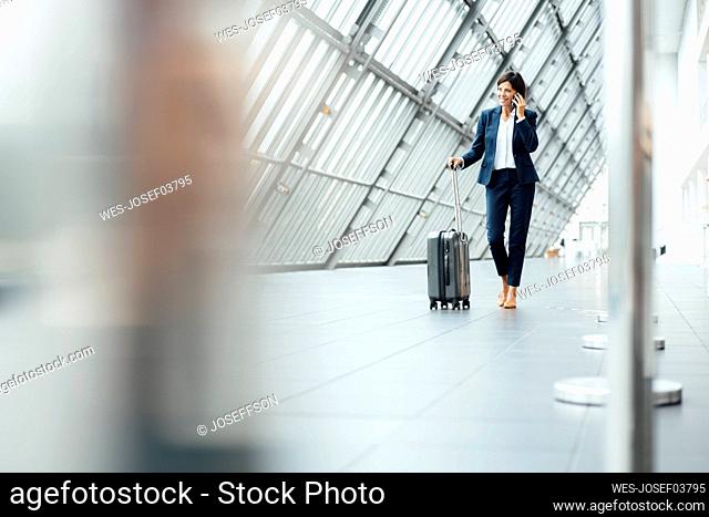 Female professional with suitcase talking on smart phone at corridor