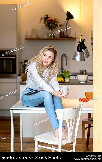 Smiling blond freelancer holding coffee cup sitting with laptop on table in kitchen at home