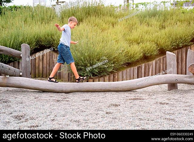 Blond boy walks on a wooden beam in a playground in a public park