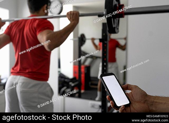 Hand of instructor holding smart phone near young man lifting barbell at gym