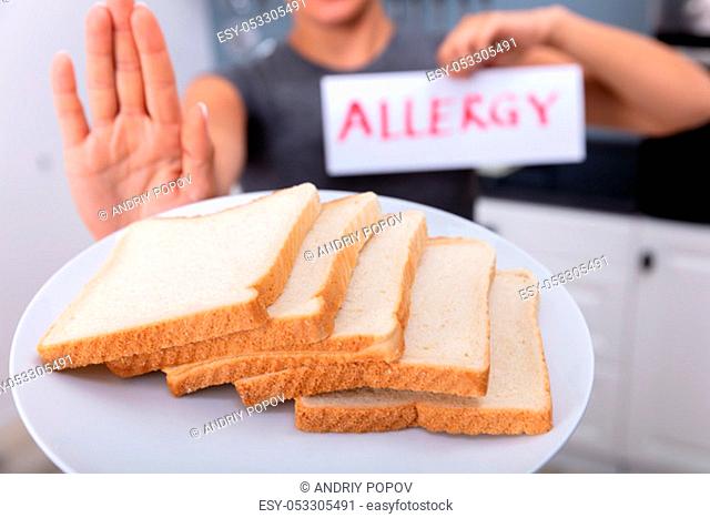 Young Woman Refusing Bread Slice On Plate At Home Holding Allergy Text Card