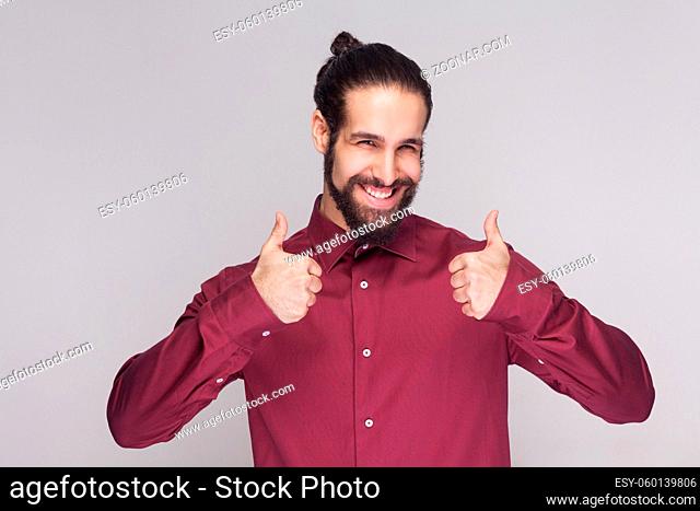 Portrait of handsome man with dark collected long hair and beard in red shirt standing, thumbs up and looking at camera satisfied, and toothy smile