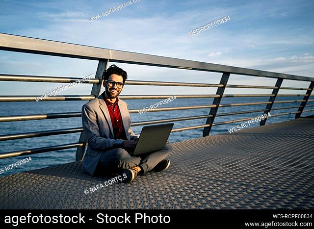 Smiling freelance worker with laptop sitting cross legged on pier