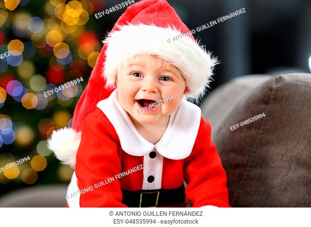 Joyful baby wearing a red santa clays disguise on a couch at home in christmas with a tree in the background