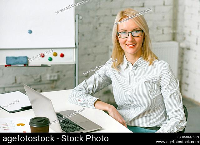 Smiling businesswoman works laptop in office. Charming blonde woman smiles looking at camera while sitting at office desk with computer on it