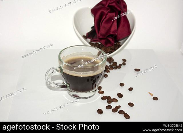 Glass cup with black drink coffee with foam floating on the liquid next to a red handkerchief and roasted coffee beans contained in a white ceramic heart