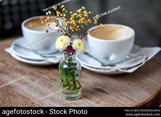 16 August 2021, Bavaria, Munich: A small vase of flowers sits on a table in a downtown sidewalk cafe in front of two used, empty coffee cups