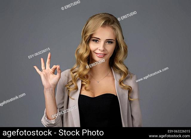 smiling young curly woman showing OK gesture on gray background