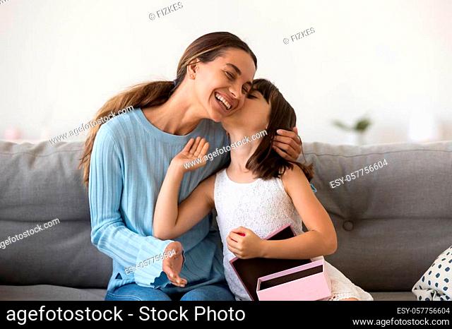 Mother daughter sitting together in living room on couch celebrating special occasion. Adorable grateful sincere child kisses mother received surprise pink gift...