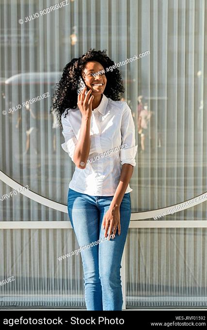 Smiling businesswoman talking on mobile phone while standing in front of glass wall