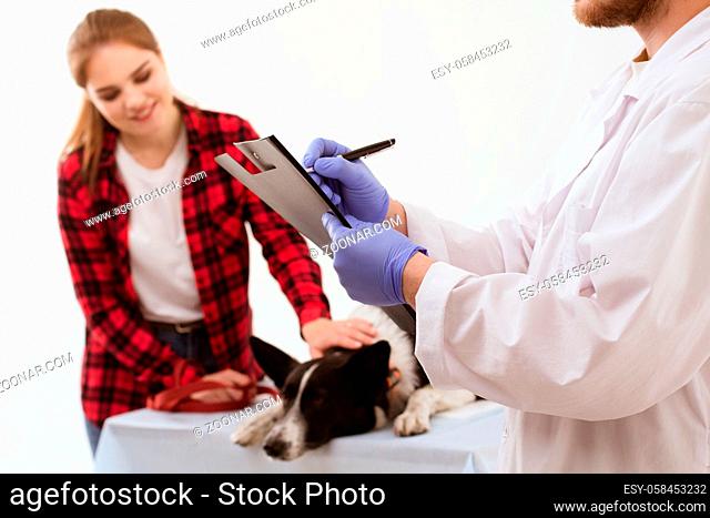 Male vet writing on clipboard while his patient waits in background. Pretty woman petting her dog at vets appointment while doctor fills in certain information...