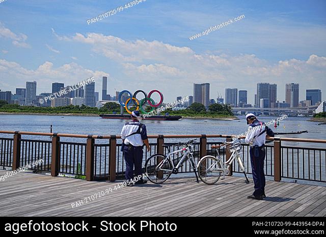 20 July 2021, Japan, Tokio: Two security personnel stand on their bicycles on the shore in front of the Olympic rings. The rings are on a pontoon in the bay of...