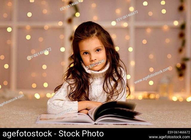 Happy little child girl with fairy tale book looking at the camera on the background with lights. Merry Christmas. space for text
