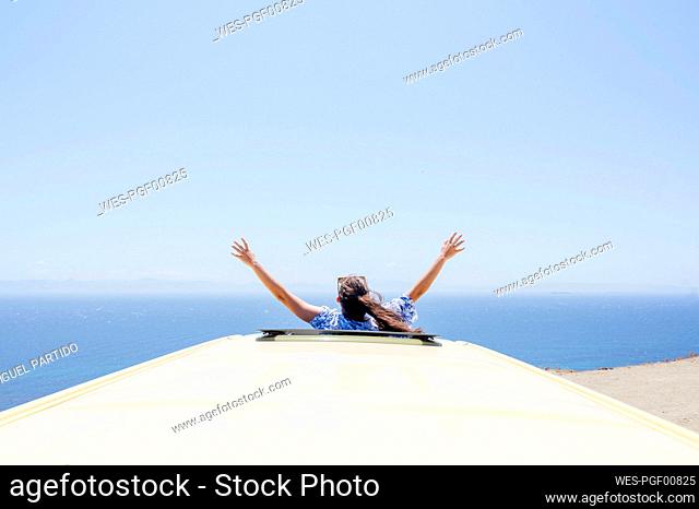 Woman with arms raised leaning through sunroof of van during sunny day