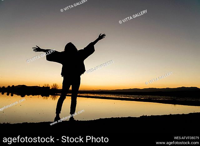 Young woman with arms outstretched standing by river during dusk at Ebro delta, Spain