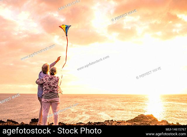 close up and portrait of two old and mature people playing and enjoying with a flaying kite at the beach with the sea at the background with sunset - active...