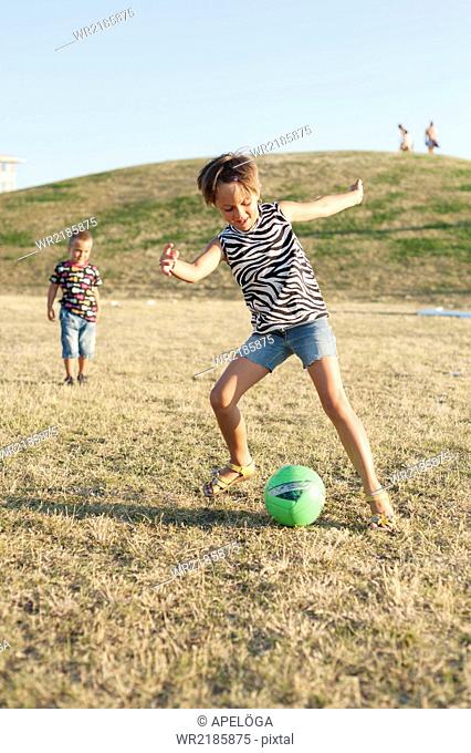 Little girl kicking ball while brother looking at her on grassy field