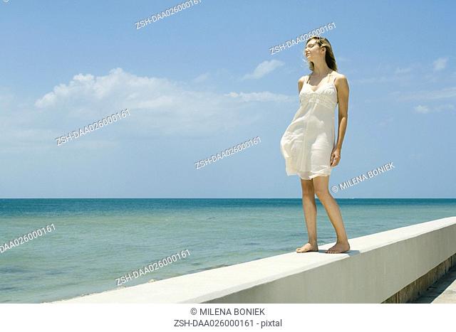Young woman in sundress standing on low wall beside sea, eyes closed