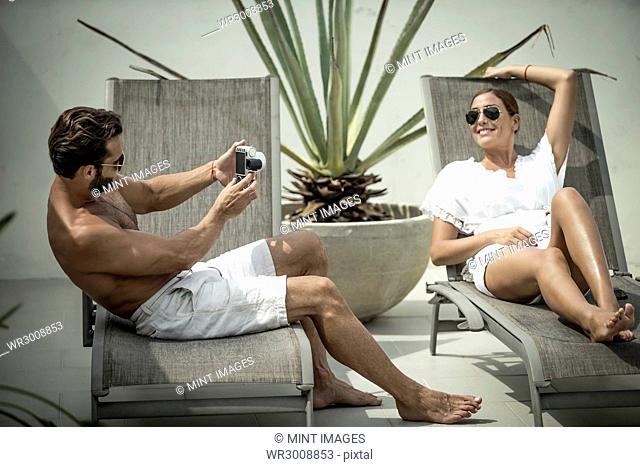 A seated man taking a picture of a woman lying on a sun lounger