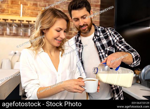 Handsome young husband adding milk or coffee creamer to his girlfriend cup enjoying breakfast together in kitchenn at home