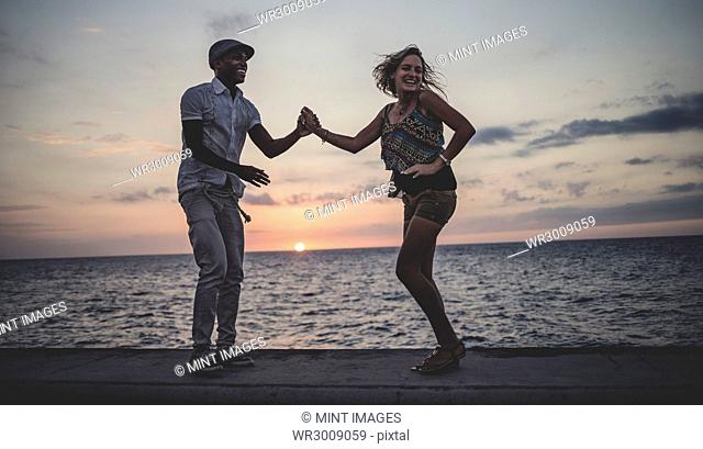 Two people dancing on a sea wall in front of the ocean at dusk