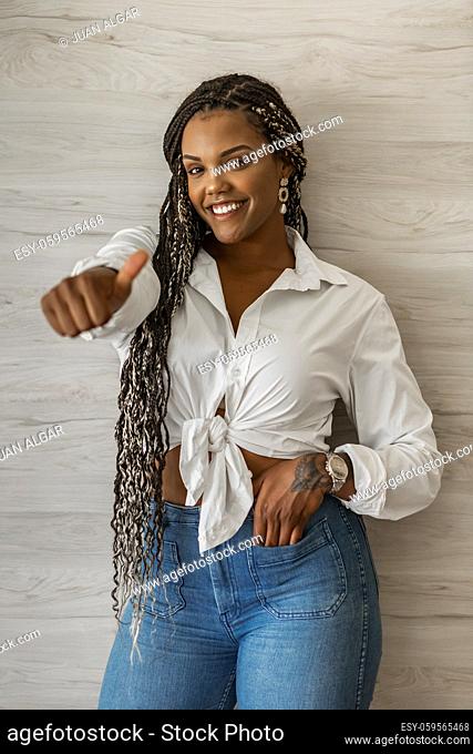 Portrait of a smiling African American adult woman standing doing thumb up and looking at the camera. Adult woman with black and yellow braids posing on a wall...
