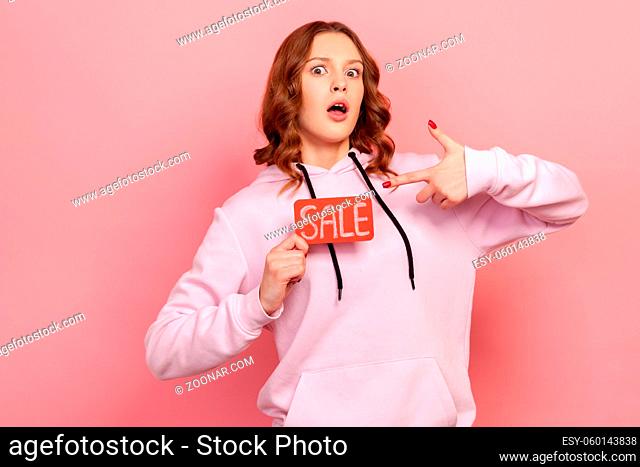 Portrait of surprised curly haired teenage girl holding sale word, opening mouth in amazement, looking shocked by discounts