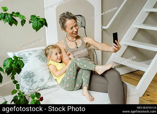 Smiling woman taking selfie with girl through smart phone at home