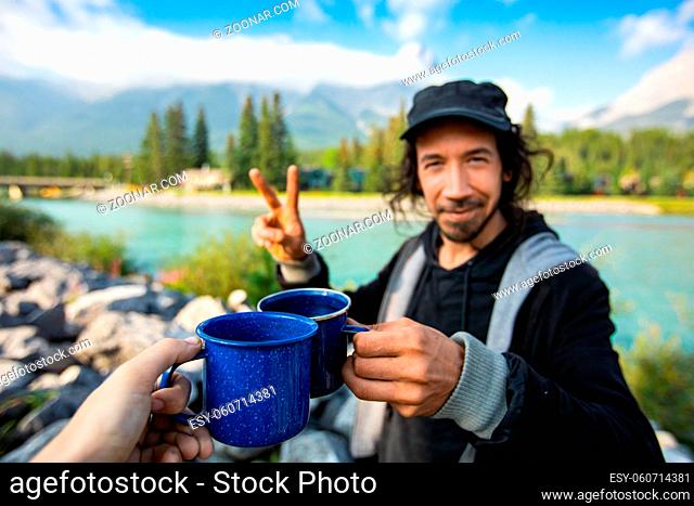 Selective focus of young middle aged couple drinking coffee and tea while standing in front of a blue river and mountains in the background