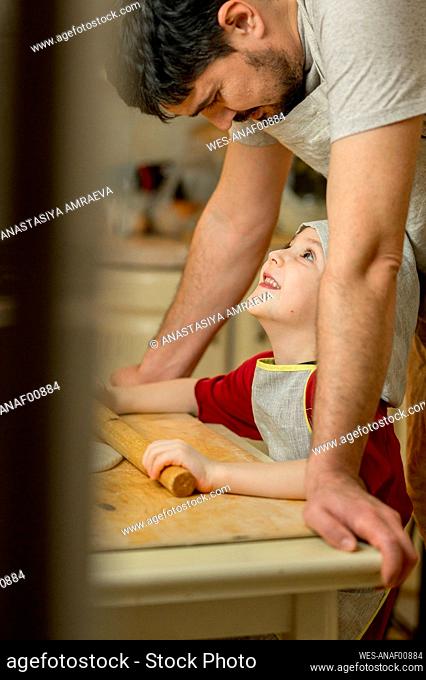 Smiling boy with rolling pin looking at father and preparing food in kitchen