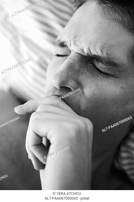 Man's face with eyes closed and furrowed brow, hand over mouth, lying on bed, close-up, black and white