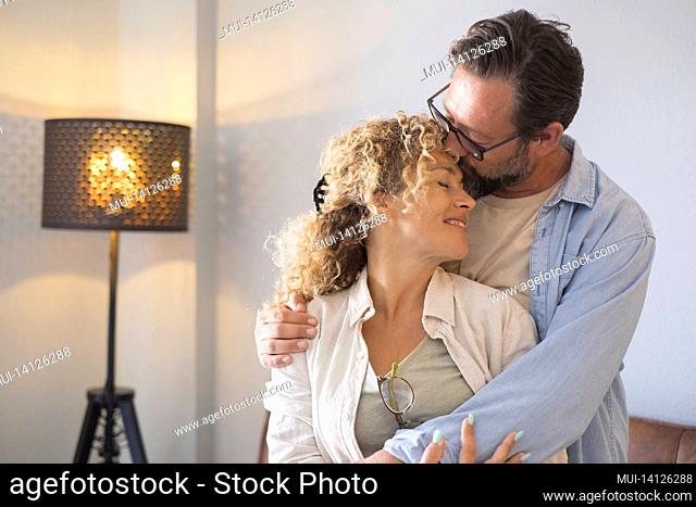 couple embracing and romancing standing in living room at home. husband kissing forehead of wife with affection. affectionate couple spending leisure time...