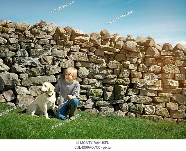 Boy and dog sitting by dry stone wall in field