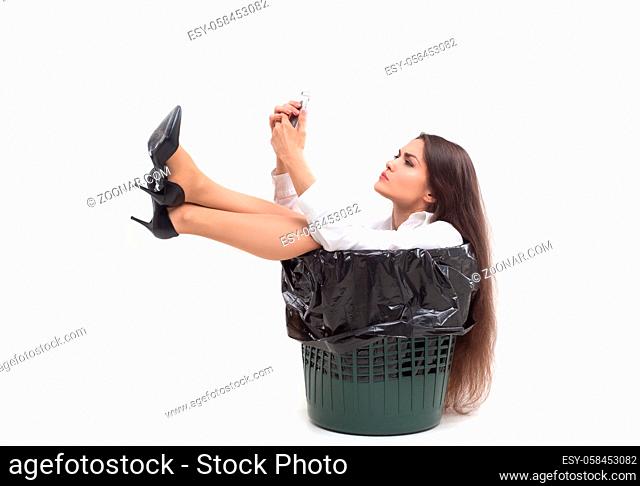Studio shot of female sitting in garbage can. Crazy female sits in bucket ment for garbage with her legs out looking at her phone she is holding in her hands