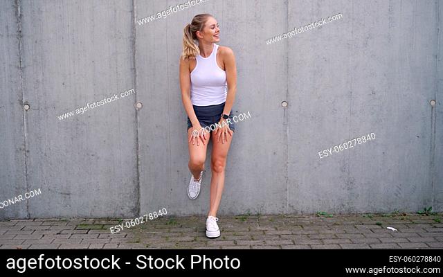 Full body delighted young sportswoman in shorts and top leaning on concrete wall and looking away with smile while resting during fitness training on street