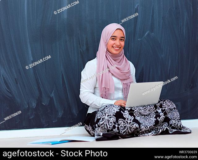 arab female student working on laptop from home black chalkboard in background