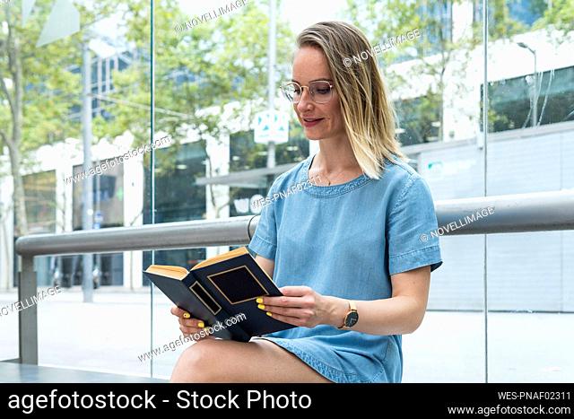 Businesswoman reading book while sitting in front of glass wall