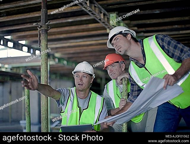 Male construction worker pointing while discussing with coworkers over blueprint at construction site