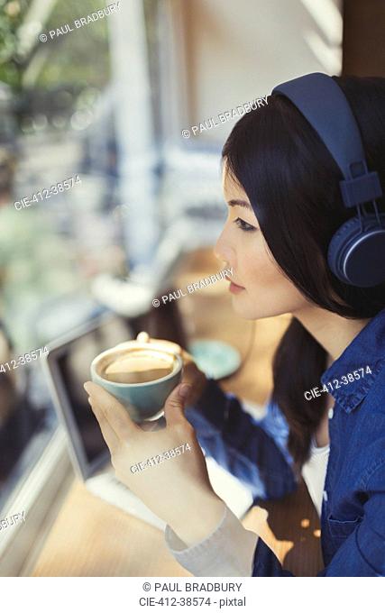 Pensive young woman drinking coffee, listening to music with headphones at cafe window
