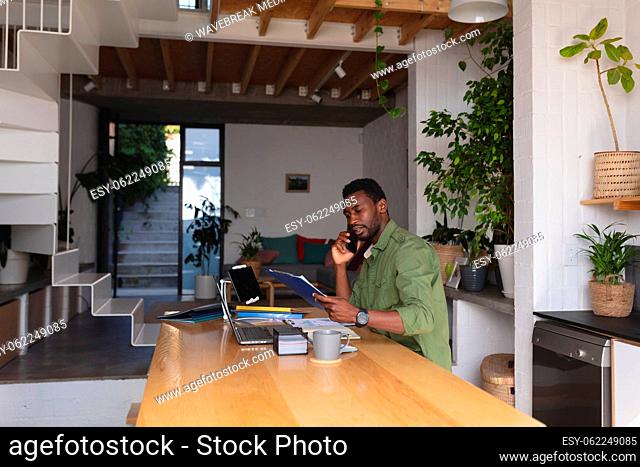Happy african american man sitting at table in kitchen using laptop and talking on smartphone