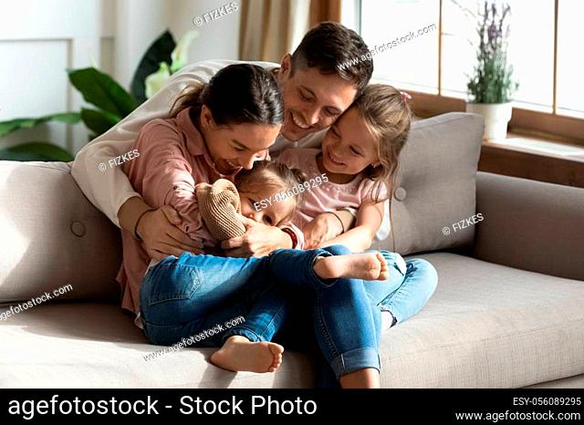 Happy cheerful parents having fun with cute children daughters cuddling playing on sofa together, mom and dad laughing embracing little kids daughters tickling...