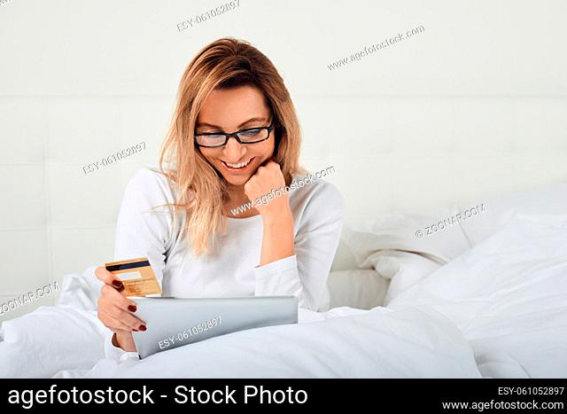 Attractive woman relaxing in bed doing online shopping holding her credit card in one hand and digital tablet in the other as she smiles at the camera